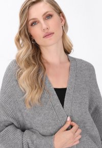Grå stickad cardigans med V-ringning, löst sittande och ribbad textur. Modellen har på sig en svart topp under, med axellångt vågigt hår.