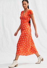 Abito rosso a pois con gonna ampia, maniche corte a volant e un nodo in vita. Indossato con stivali bianchi alla caviglia con un bordo decorativo.