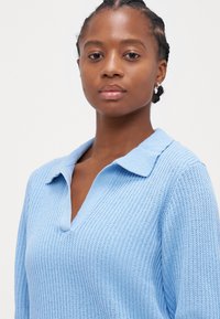 Suéter de pulôver em malha azul claro com colarinho, apresentando um padrão texturizado e um decote em V. Mangas curtas e ajuste descontraído.