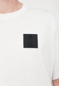 Hvit bomull t-skjorte med et kvadratisk svart lærpatch med preget logo og teksten "JUST CAVALLI" på venstre side.