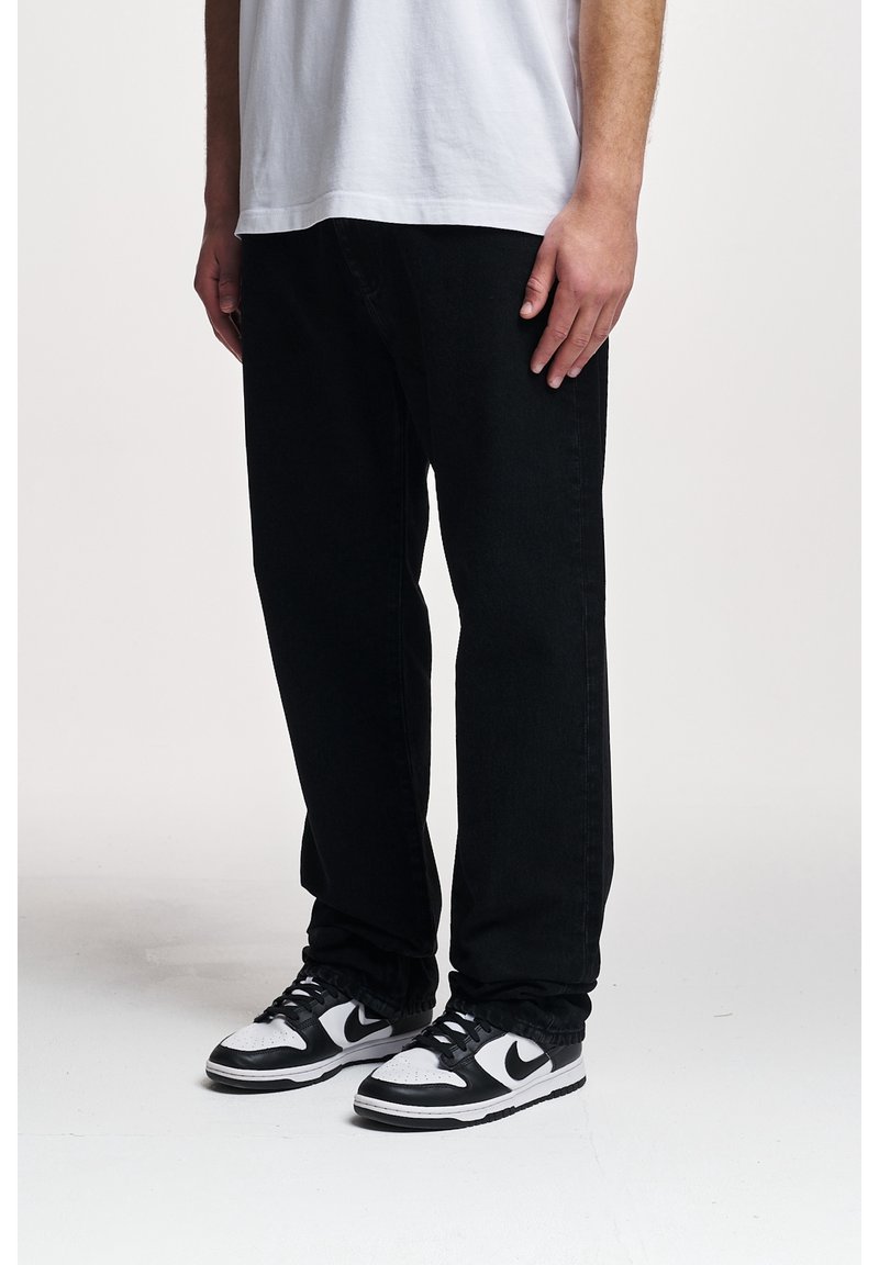 Partie inférieure du corps d'une personne portant un pantalon noir ample, des baskets noires et blanches et une chemise blanche sur un fond uni.