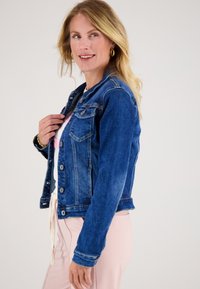 Blonde vrouw die gouden oorringen draagt, een blauwe denimjas over een wit shirt en lichtroze broek, glimlacht en naar de zijkant kijkt.