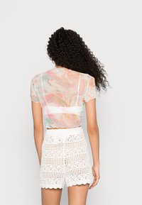 Top cropped transparente com um padrão marmorizado colorido em rosa, verde e branco; combinado com calções brancos de crochê com bordas onduladas.