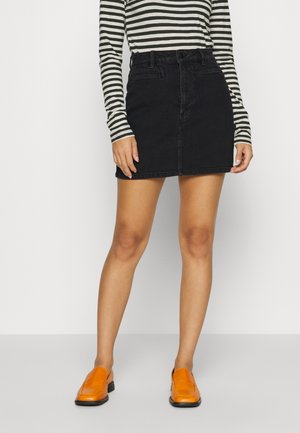 Femme portant une mini-jupe en denim noire, un haut à manches longues rayé noir et blanc, et des mocassins orange vif, debout devant un fond uni.