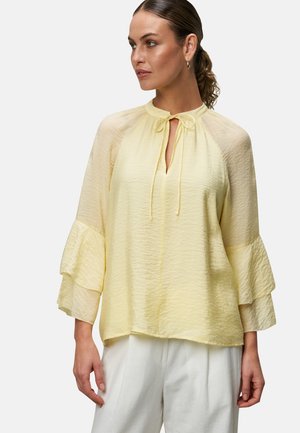 Blouse jaune avec un détail en trou de serrure et une fermeture par nœud à l'avant. Présente des manches transparentes à volants et un tissu léger et texturé.