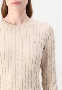 Pulover tricotat cu rețea bej, având un design cu cabluri și un guler rotund. Include un mic logo brodat pe piept.