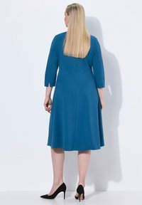 Turkusowa sukienka do kolan z 3/4 rękawami i bocznymi rozcięciami, wykonana z gładkiego materiału. Połączona z czarnymi szpilkami.