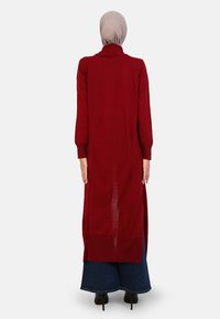 Long burgundy cardigan with a high collar, full-length sleeves, side slits, and a loose fit, paired with wide-leg denim trousers.