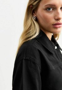 Chaqueta de mezclilla negra con un gran cuello, cierre a presión y detalles de costura visibles. El modelo tiene el cabello largo y se muestra de perfil.