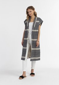 Gilet lungo, aperto davanti, con un motivo bianco e nero realizzato in tessuto intrecciato, caratterizzato da maniche corte e spacchi laterali, abbinato a una maglietta bianca e pantaloni.