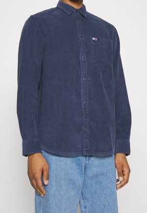 Man wearing navy blue corduroy button-up shirt with chest pocket and light blue jeans, facing forward with arms relaxed.