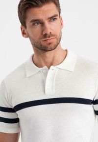 Vits kortärmad polo med ribbad textur och marinblå vågräta ränder över bröstet. Har en tvåknapps placket och krage.