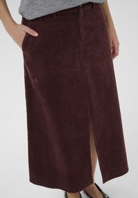 Gonna in corduroy bordeaux con righe verticali, spacco fino al ginocchio e tasca laterale. Presenta una vita con chiusura a bottone.