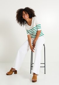 Camisa blanca a rayas con líneas verdes y beiges, de escote en V y mangas cortas. Combinada con pantalones acampanados blancos y sandalias de tacón abiertas de dedo en marrón.