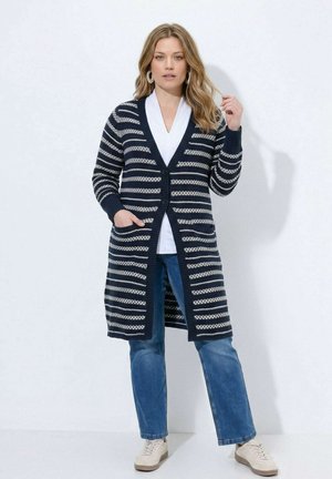 Femme portant un long cardigan rayé bleu marine et blanc, un jean bleu, une chemise blanche et des baskets beige, debout sur un fond blanc uni.