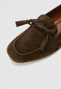 Scarpa mocassino in suede marrone con punta rotonda, caratterizzata da un dettaglio a fiocco con frange e cuciture a contrasto lungo il bordo superiore.