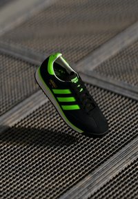 Black sneaker with vibrant green accents, featuring three stripes, textured fabric, and a retro design; lightweight and streamlined shape.