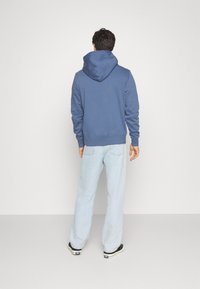 Felpa blu con cappuccio regolabile, orlo e polsi a coste, abbinata a jeans chiari in denim e sneakers nere. Vista da dietro.