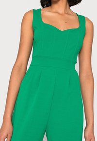 Green jumpsuit with a fitted bodice, sweetheart neckline, and wide straps. Features pleats at the hips for added texture.