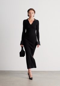 Vestido preto de manga longa com texturização em ribanas, apresentando um decote em V, detalhes de ombros decorativos com tachas e punhos expandido. Modelo segurando uma pequena bolsa preta.