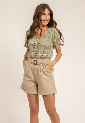 Femme souriante, portant un haut en maille à manches courtes et un short beige taille haute avec une ceinture, debout les mains dans les poches.