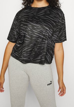 Frau trägt ein schwarzes und graues, locker sitzendes T-Shirt mit Zebramuster und hellgraue Puma-Leggings, steht mit entspannt an den Seiten hängenden Armen.