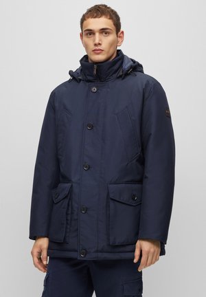 Navy blue padded jacket with a high collar, hood, and front-button closure. Features two large pockets and a smooth, matte finish.