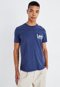 Granatowy bawełniany t-shirt z krótkimi rękawami, okrągłym dekoltem i białym nadrukowanym logo po lewej stronie klatki piersiowej. Luźny krój, miękka tekstura.