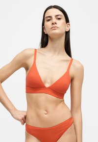 Femme aux longs cheveux foncés portant un soutien-gorge orange et une lingerie assortie, debout devant un fond blanc uni.