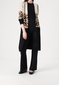 Długi kardigan w kolorach kremowym, karmelowym i czarnym. Dziergany materiał z motywami zwierzęcymi. Otwarty przód, ściągacze na mankietach i dole. Noszony na czarnym topie i spodniach.