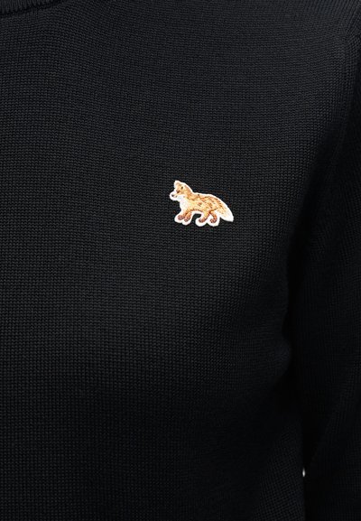 Μικρό κεντητό πατς με πορτοκαλί και λευκή αλεπού σε μαύρο πλεκτό πουλόβερ, τοποθετημένο κοντά στην περιοχή του στήθους.
