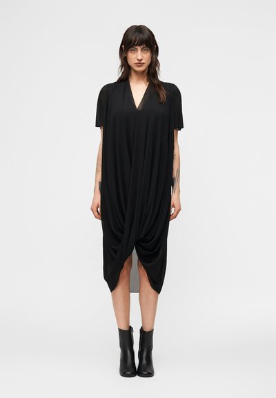 Femme portant une robe noire drapée longueur genou avec des manches courtes et des bottines noires, debout devant un fond clair uni.