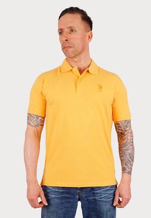 Polo shirt orange en coton avec des manches courtes et un col. Présente un petit logo brodé sur la poitrine. Porté avec un jean bleu.