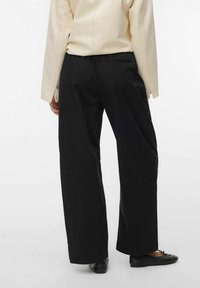 Personne portant un pantalon noir à jambes larges, un haut beige à manches longues et des ballerines noires, debout devant un fond blanc uni.