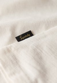 Hvidt stof med en blød tekstur, der har et sort mærke med inskriptionen "Superdry" i guld. Stofoverfladen viser fine, lette ribbede strukturer.