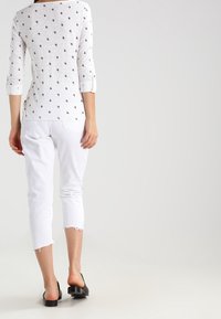 Weißes Oberteil mit 3/4-Ärmeln und schwarzem Tropfenmuster, kombiniert mit weißen, knöchellangen Jeans mit fransigen Säumen und schwarzen flachen Schuhen.