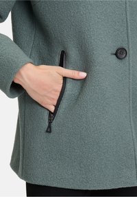 Grøn uldblandet frakke med en blød tekstur, der har en sort lynlåslomme og en enkelt sort knaplukning.