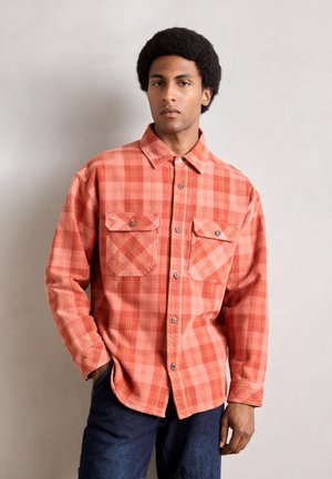 Plaid shirt in orange tones, button-up design with two chest pockets, long sleeves, and rounded hem. Fabric appears soft with a textured finish.