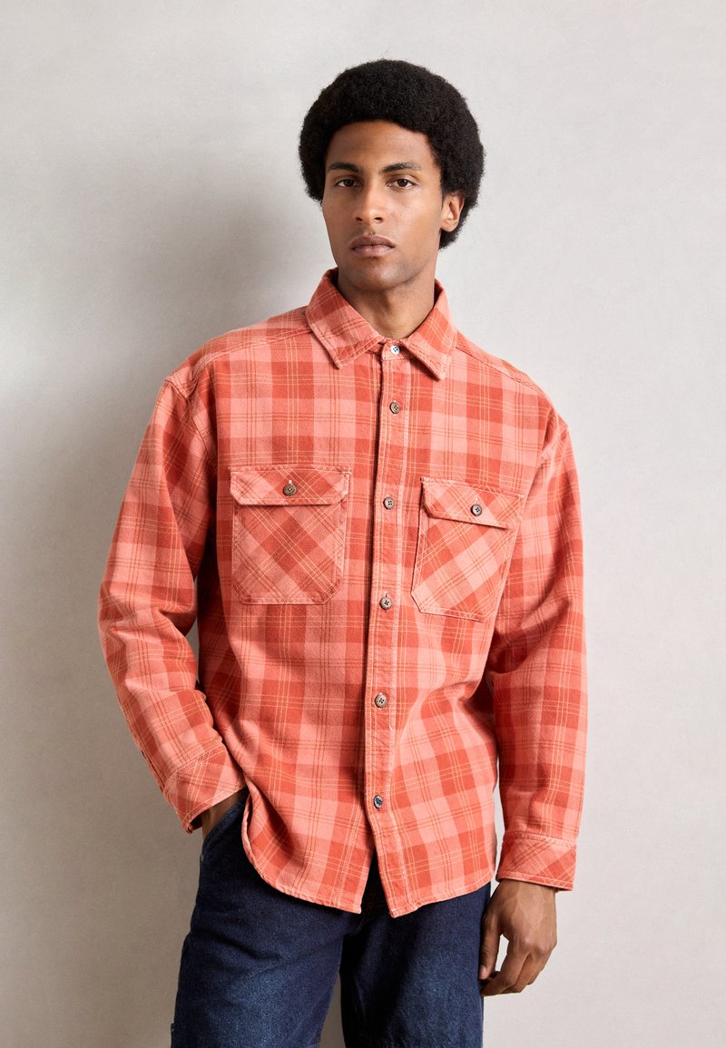 Camisa de cuadros en tonos naranjas, diseño de botones con dos bolsillos en el pecho, mangas largas y dobladillo redondeado. La tela parece suave con un acabado texturizado.