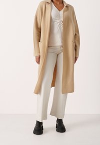 Beige wollen jas met grote zakken, lange mouwen en een ontspannen pasvorm, gedragen over een witte top en gecombineerd met crèmekleurige broeken en zwarte stevige laarzen.