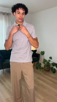 Jonge man die de kraag van een lichtgrijs poloshirt rechtzet, staand in de woonkamer met beige broek, groene bank en potplanten op de achtergrond.