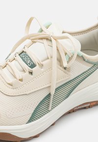 Scarpa sportiva beige con parte superiore in mesh texturizzato, dettagli verdi e lacci crema. Presenta una suola con motivo e il marchio "PUMAGRIP ATR".