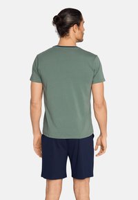 Mies, jolla on tummat hiukset nutturalla, pukeutuneena lyhythihaiseen vihreään T-paitaan ja tummansinisiin shortseihin, seisoo selin katselijaan.