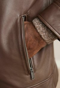 Barna bőrkabát cipzáras zsebbel, amelyen a nadrágszáron látható egy kötött mandzsetta. Sima textúra és testhezálló fazon.