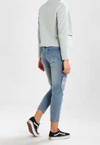 Světle šedá zkrácená bunda s texturovaným povrchem, modré distressed džíny s nášivkou a černobílé tenisky.
