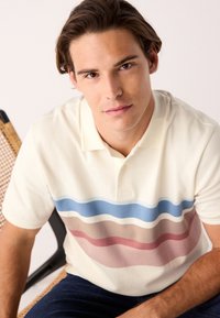 Polo bianca con strisce orizzontali in blu, rosa e beige. Maniche corte, colletto e tessuto strutturato.