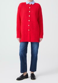Red ribbed cardigan with large golden buttons, layered over a blue and white striped collared shirt. Paired with blue denim pants and black loafers.