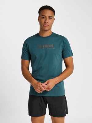 Jonge man staand, draagt een turquoise "hummel performance" t-shirt en zwarte korte broek, handen voor zich geklemd, neutrale uitdrukking, witte achtergrond.