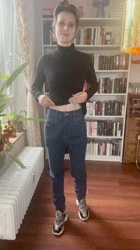 Top de gola alta preto, jeans de ganga azul de cintura alta com corte reto e ténis estampados. O fundo apresenta prateleiras de livros e plantas.