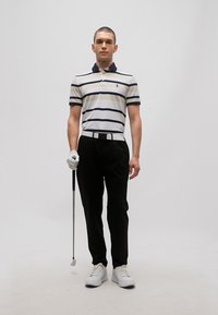 Jeune homme debout tenant un club de golf, portant un polo rayé, un pantalon noir, un gant de golf blanc à la main gauche, une ceinture blanche et des baskets blanches.
